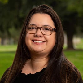 UCR Staffer Sarah Cleary. Sarah has dark brown hair that goes below her shoulders. She has a smile on her face, wears glasses and a dark shirt. The background has green grass and trees.