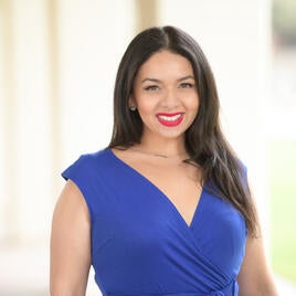 UCR staffer Cristina Gonzalez. She has dark hair that is pulled behind her. Cristina wears a bright smile with red lipstick and a ribbed blue shirt. The background has golden leaves.